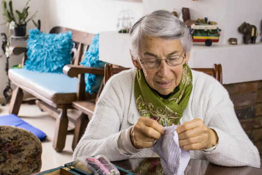 An older woman wearing a green scarf with a fabric face mask in her hands and sewing supplies on the table beside her.