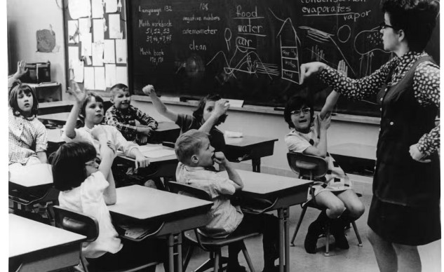 A white woman stands by a classroom blackboard in front of white students sitting at desks, many with their hands raised.