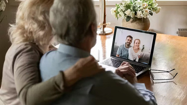 A caregiver sitting at a desk, looking at a laptop while holding a phone, symbolizing the challenges of coordinating care remotely from a long distance.