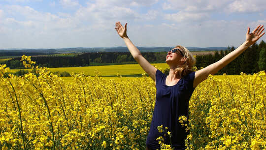Can Meditation Be Fun? Or Does It Have To Be Serious women standing in a field of flowers, smiling with her hands open up to the sky