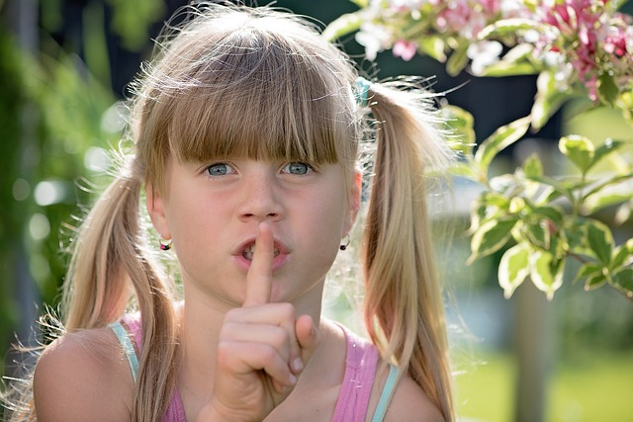 a young girl with her finger over her mouth in the universal symbol of "hush, be quiet"