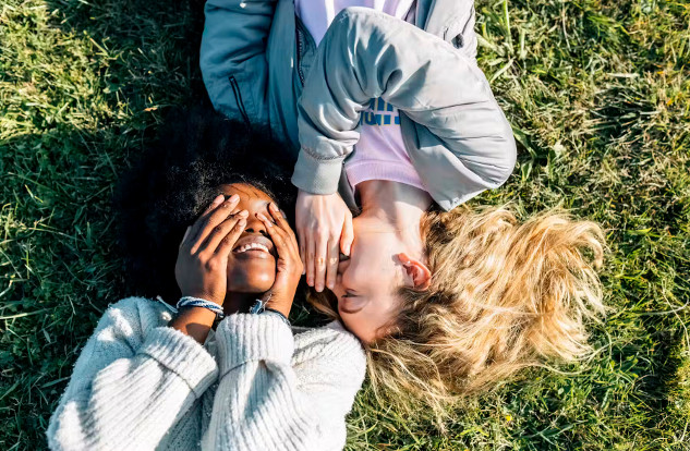 Two smiling people lying on grass, one with hands over eyes and the other whispering into their ear. 